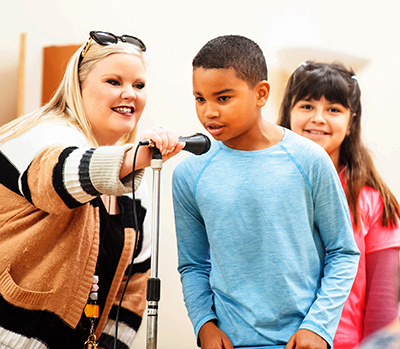 boy standing at microphone held by teacher